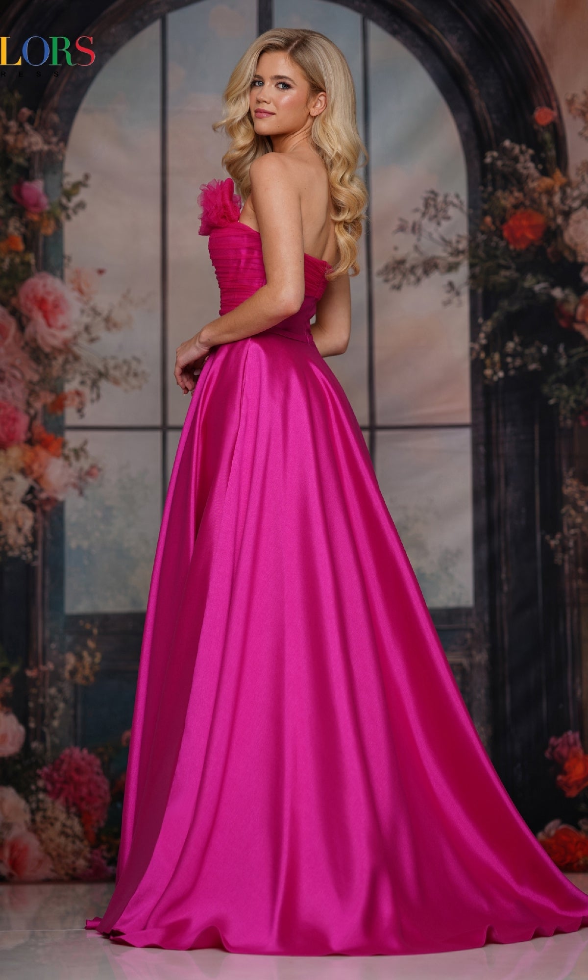 A woman in the Strapless Long A-Line Prom Dress: Colors Dress 3737 stands before a floral, arched window backdrop, glancing over her shoulder in a bright pink gown.